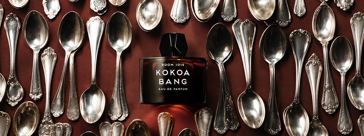 Kokoa Bang de Room 1015, eau de parfum, flacon posé sur du velours bordeaux parmi des cuillères en argent anciennes.