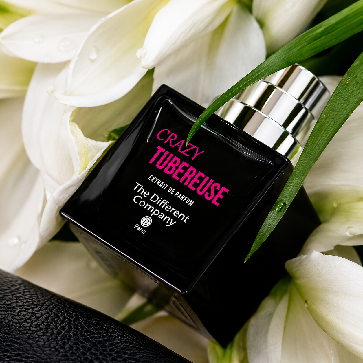 Crazy Tubéreuse de The Different Company, extrait de parfum, flacon en mise en scène sur du cuir noir, entouré de fleurs de tubéreuse blanches.