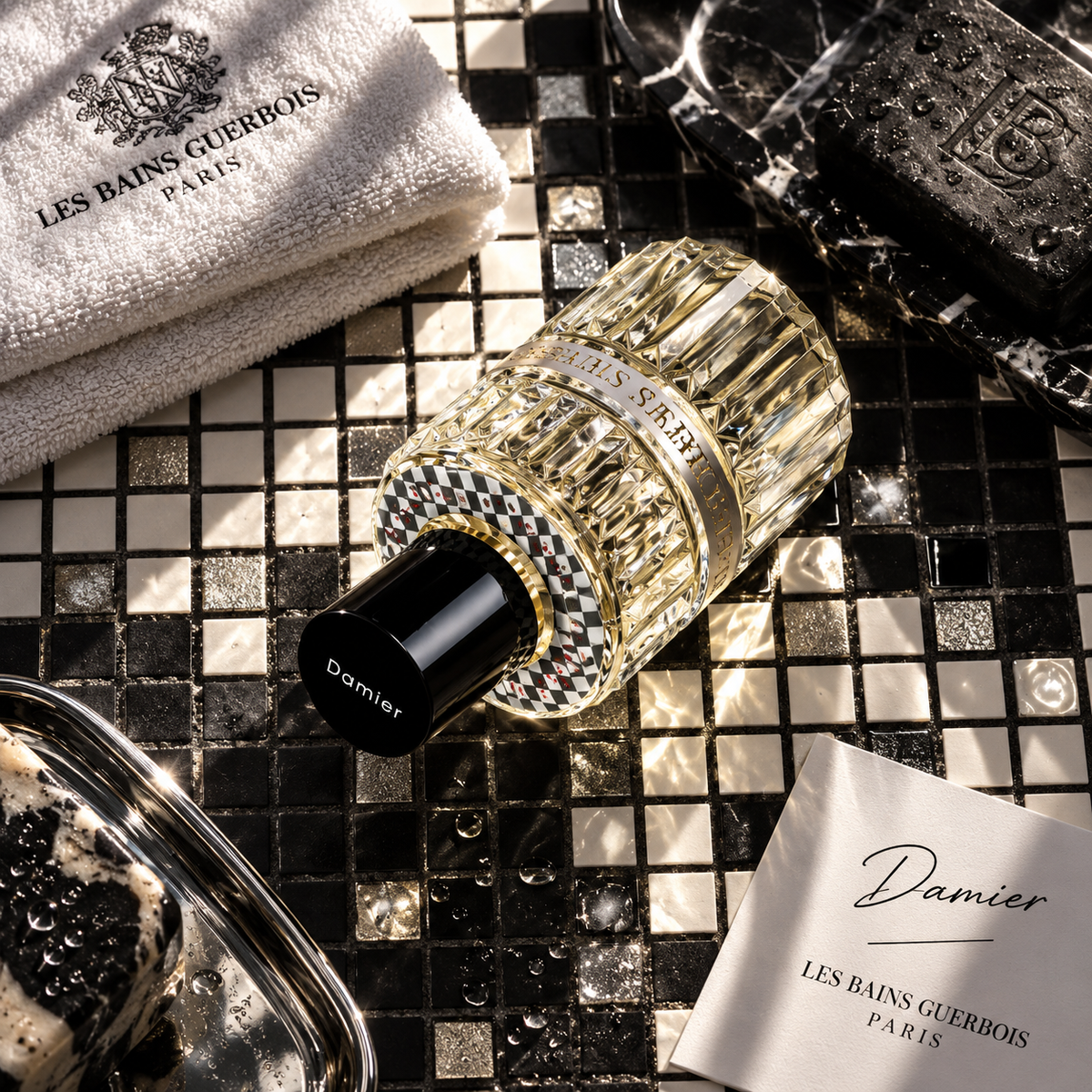Damier de Les Bains Guerbois, eau de parfum 100 ml, flacon couché sur une mosaïque noire et dorée, serviette et savon de la maison.