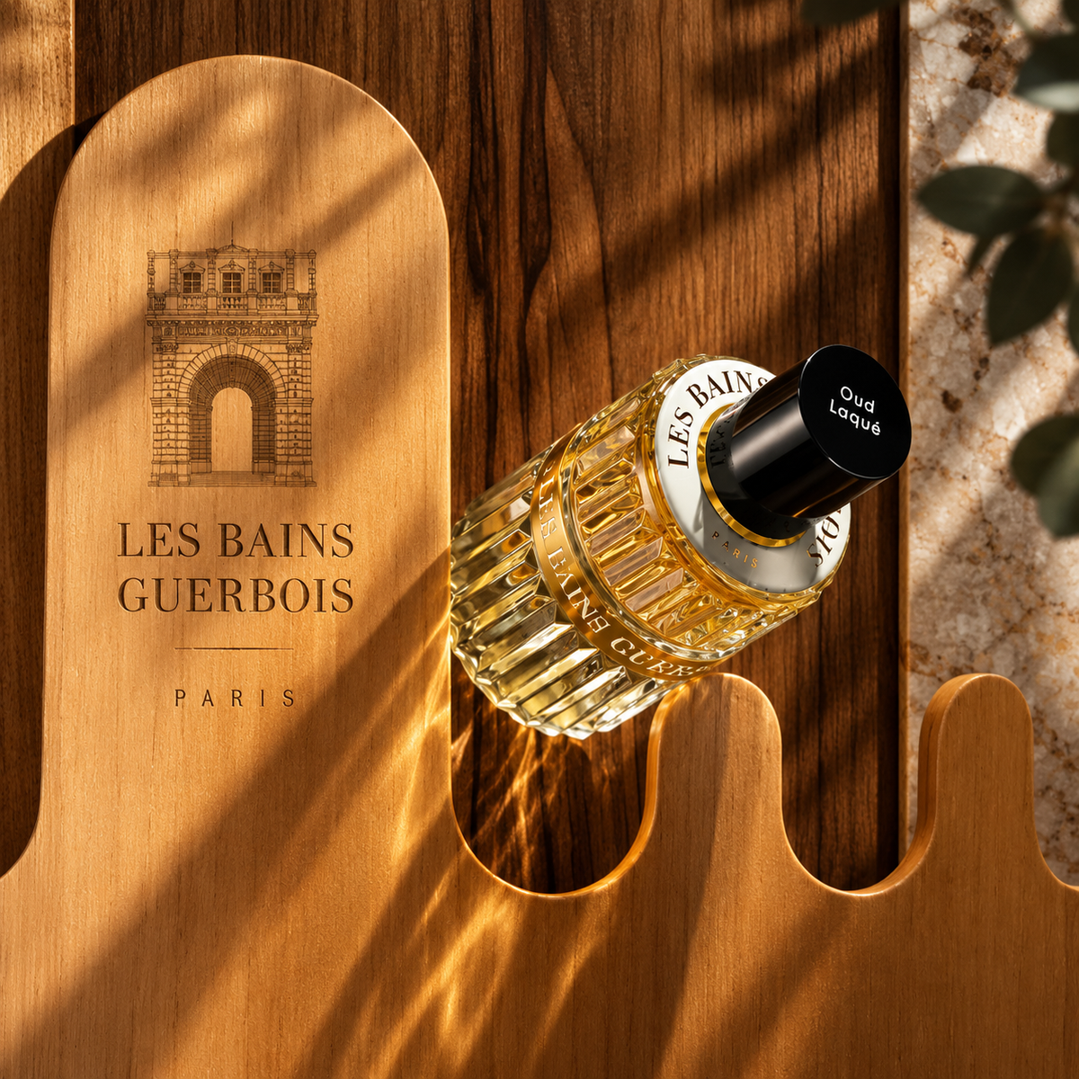 Oud Laqué de Les Bains Guerbois, eau de parfum, flacon incliné sur un panneau de bois clair gravé au nom de la maison.